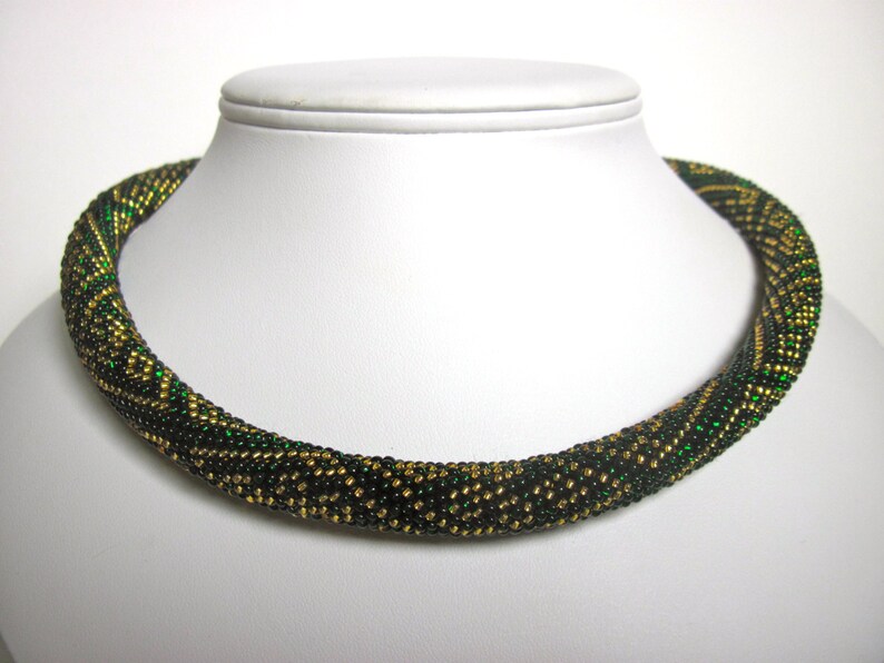 Op de afbeelding: Een groene en gouden kralenketting met een geometrisch patroon. De ketting is gemaakt van kleine, ronde kralen en is ongeveer 46 cm lang.