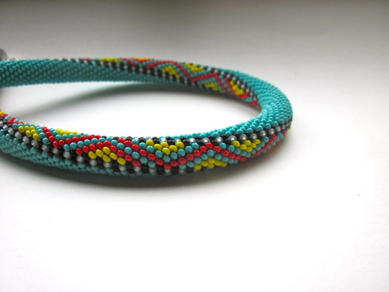 May include: Turquoise beaded bracelet with a colorful pattern. The bracelet features a repeating design of red, yellow, black, and white beads. The bracelet is made of small, round beads and has a clasp.