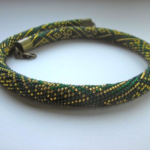 Op de afbeelding: Een groene en gouden kralenketting met een sluiting. De ketting is gemaakt van kleine, ronde kralen die in een patroon zijn geweven.