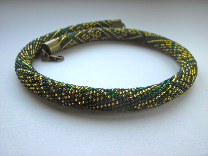 Op de afbeelding: Een groene en gouden kralenketting met een sluiting. De ketting is gemaakt van kleine, ronde kralen die in een patroon zijn geweven.