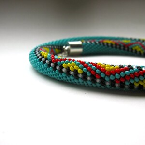 May include: A turquoise beaded bracelet with a geometric pattern in red, yellow, black, and white. The bracelet is cylindrical and has a silver clasp. The beads are small and tightly woven together, creating a textured surface.