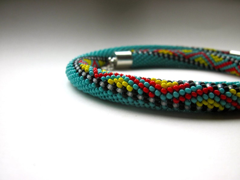 May include: A turquoise beaded bracelet with a geometric pattern in red, yellow, black, and white. The bracelet is cylindrical and has a silver clasp. The beads are small and tightly woven together, creating a textured surface.