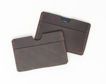 May include: Two brown leather cardholders with stitched edges. One has a curved cutout, and both feature a small embossed logo. The cardholders are displayed on a white background.