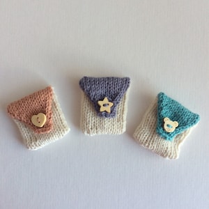 May include: Three small, hand-knitted pouches in soft pastel colors. Each pouch features a flap and a wooden button closure. One has a heart button, another a star, and the last a bear. The pouches are set against a white background.