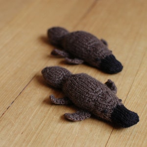 May include: Two knitted platypus toys, brown with black beaks and tails. They are lying on a light brown wooden surface. The toys are handmade.