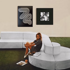 May include: A white modular sofa with a curved design, featuring a woman sitting on the sofa wearing a black and white polka dot dress. Two black framed prints hang on the wall behind the sofa. One print features a black and white geometric design, and the other print features a black and white design of overlapping circles.