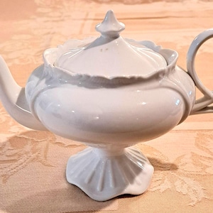 May include: A white porcelain teapot featuring a rounded body, a curved spout, and a looped handle. The lid is topped with a pointed finial, and the base is pedestal-style with a scalloped edge. The rim of the teapot body also has a scalloped design.