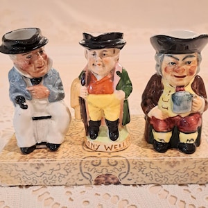May include: Three ceramic character figurines standing on a rectangular base. The left figure wears a blue coat and white trousers. The center figure, labeled Tony Weller, wears a green coat and yellow breeches. The right figure wears a brown coat.