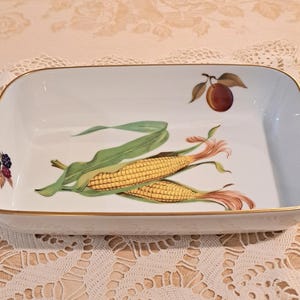 May include: A rectangular white ceramic baking dish with a gold-trimmed rim. The interior features a central illustration of two ears of yellow corn with green husks, a single peach in the upper right corner, and a cluster of blackberries on the left.