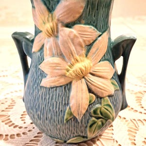 May include: A ceramic vase with a textured blue surface featuring two prominent raised light pink flowers with yellow centers and green leaves. The vessel has two symmetrical handles on the sides and a wide, open top rim with a smooth finish.