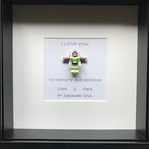May include: A framed picture with a Buzz Lightyear figure at the centre. The white background has the text "I LOVE YOU" and "TO INFINITY AND BEYOND". Below the text are the names "Clare & Steve" and the date "7th September 2021". The frame is black.