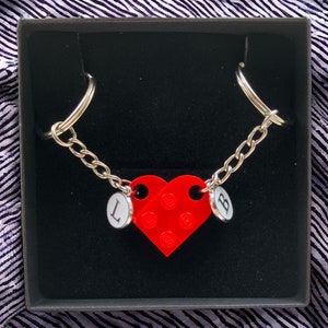 May include: A red heart-shaped keychain with the letters "L" and "B" on the sides. The keychain is attached to a silver chain with two silver rings.