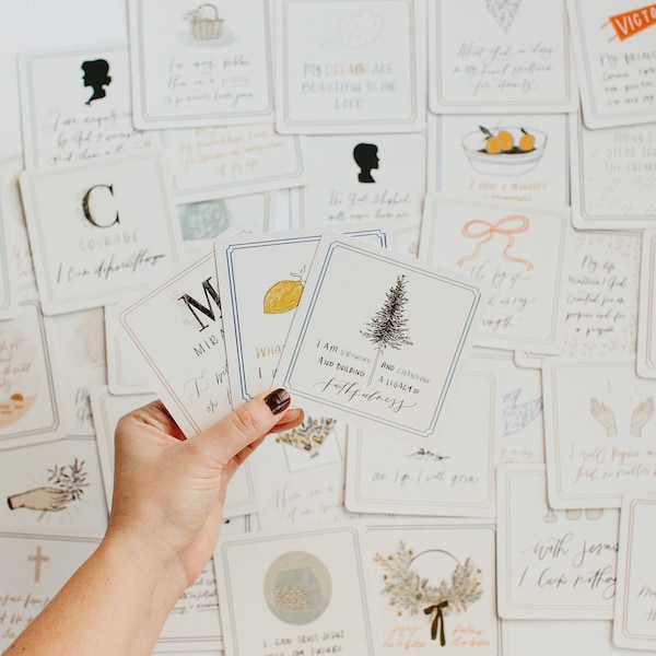 May include: A hand holds three white cards with illustrations and text, including a lemon, a pine tree, and the words "I am growing and cultivating a legacy of faithfulness." Other cards with illustrations and script are scattered in the background.