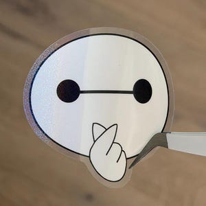May include: A white sticker featuring a cartoon character with a round head, two black eyes, and a black line connecting them. The character is making a heart shape with its hand. The sticker is being held by tweezers.