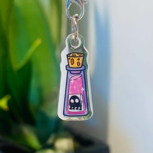 May include: A clear acrylic charm shaped like a potion bottle. The bottle features pink liquid with sparkles and a skull graphic. It has a yellow cork and a purple cap. A silver-coloured clasp is attached to the top.