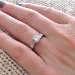 May include: A silver ring with a large, round, clear diamond in the center and smaller diamonds set in a band around the center stone.
