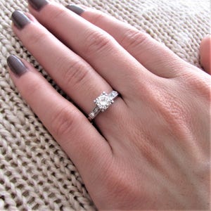 May include: A silver engagement ring with a round, clear center stone and smaller stones set in a band.