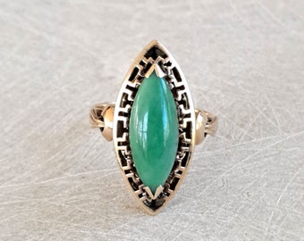 May include: A gold-toned ring featuring a large, oval-shaped, polished green stone. The stone is set within an openwork frame with a geometric design. The ring has a detailed band with decorative elements on the shoulders.