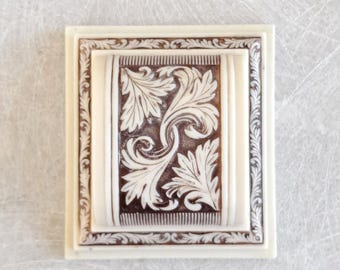 Könnte beinhalten: Rechteckige, cremefarbene Zierplatte mit detailliertem, erhabenem Blumendesign. Die zentrale Tafel zeigt einen dunkelbraunen Hintergrund mit weißen, wirbelnden Blättern. Die Platte ist mit einem sich wiederholenden Blättermuster und einem erhabenen Rand umrahmt.