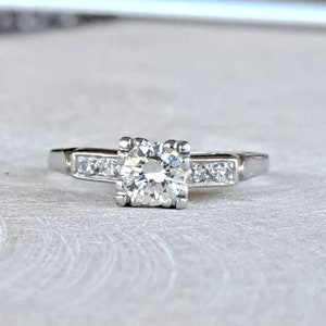May include: A silver engagement ring with a single round diamond in a square setting and smaller diamonds set in a band.
