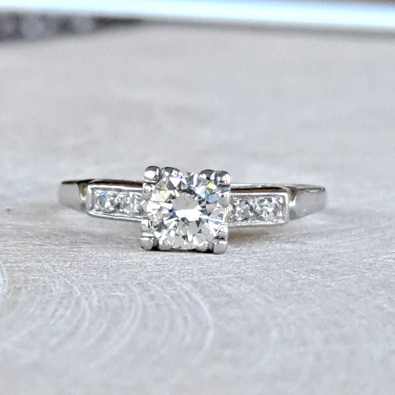 May include: A silver engagement ring with a single round diamond in a square setting and smaller diamonds set in a band.