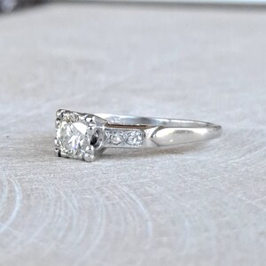 May include: A silver engagement ring with a round, clear center stone surrounded by smaller stones. The band is plain and has a slight curve.