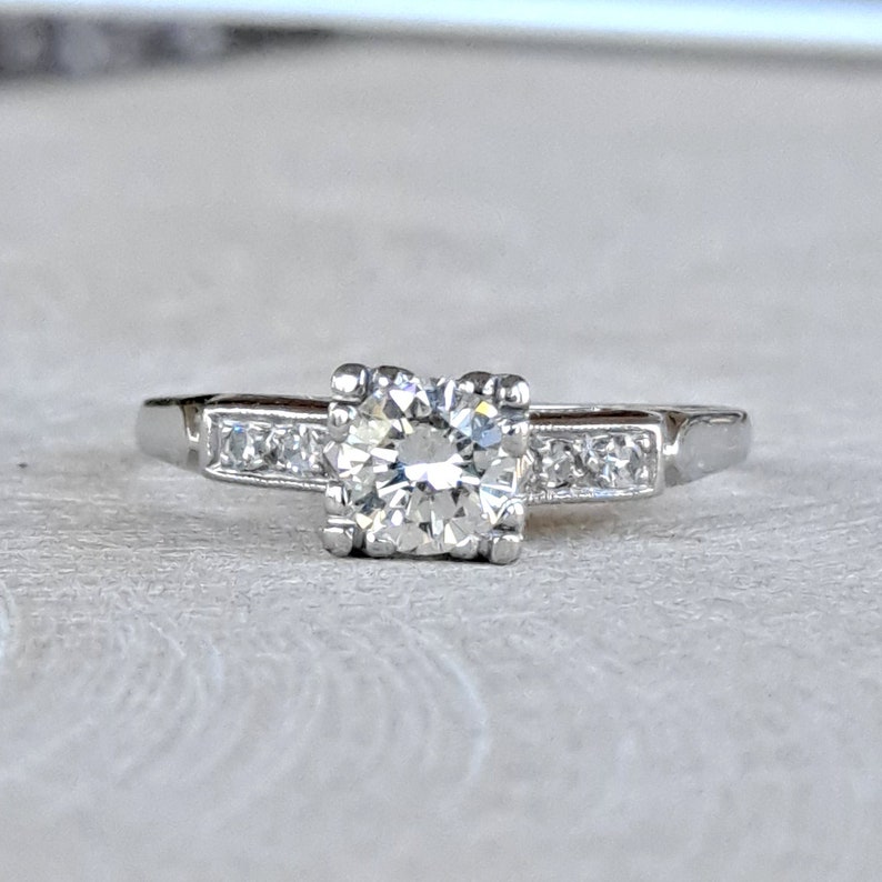 May include: A silver engagement ring with a single round diamond in the center and smaller diamonds set in a band.