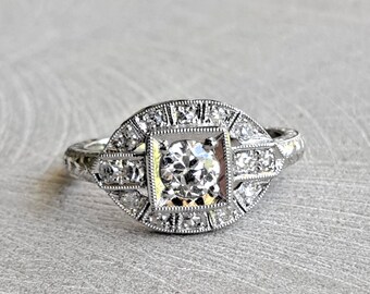 May include: A silver ring with a large, round, clear diamond surrounded by smaller diamonds in a square shape. The ring has a delicate, intricate design.