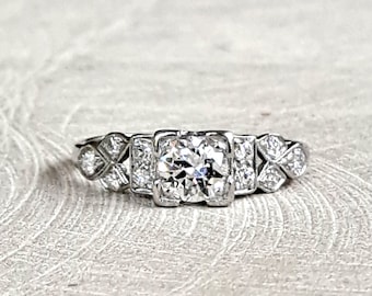 May include: A silver engagement ring with a large round diamond surrounded by smaller diamonds. The ring has a delicate, intricate design.
