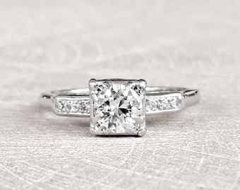May include: A silver engagement ring with a square-shaped diamond center stone and smaller diamonds set in the band.