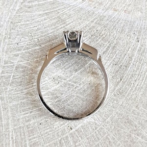 May include: A silver engagement ring with a single round diamond set in a prong setting. The band is thin and has a simple design.