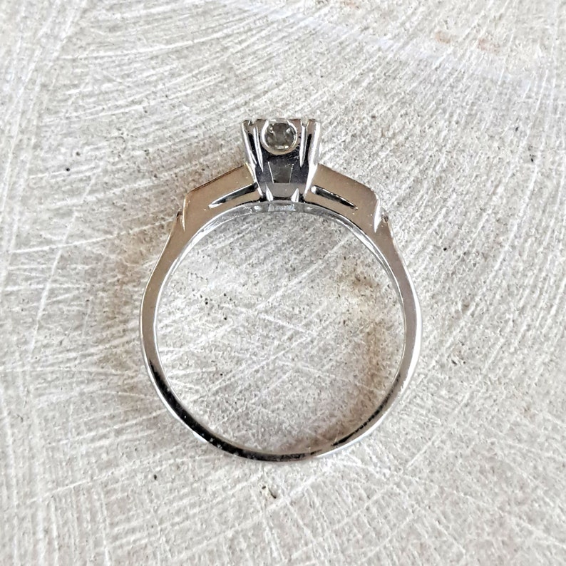 May include: A silver engagement ring with a single round diamond set in a prong setting. The band is thin and has a simple design.