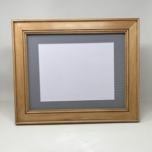May include: A rectangular picture frame with a light brown wood grain finish. The frame features a wide, flat border with beveled inner edges. Inside, a dark gray mat board surrounds a centered, blank white rectangular display area for photos.