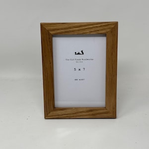May include: A rectangular wooden picture frame with a light brown finish. The frame holds a white insert with the text "The Old Plank Woodworks Est. 2013" and "5 x 7". The frame is designed to hold a 5 x 7 inch photo.