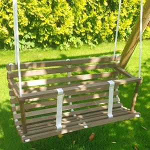 May include: A wooden swing set with a brown seat and white straps hangs from white ropes. The swing is set against a backdrop of green grass and a dense, green hedge. The swing is designed for outdoor use.