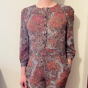 Puede incluir: Mono vintage con botones en la parte delantera y mangas largas. La prenda de una pieza tiene un estampado de cachemira y leopardo en tonos marrón, rojo y naranja. El mono tiene bolsillos y cintura entallada.