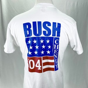Può includere: Maglietta bianca con una grafica blu e rossa di una bandiera americana con il testo "Bush Cheney '04" stampato sul retro.