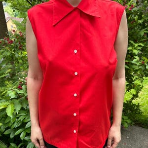 May include: A sleeveless, bright red button-down shirt with a pointed collar. The shirt has white buttons down the front and is made of a smooth fabric. The shirt is a solid color and is a classic style.