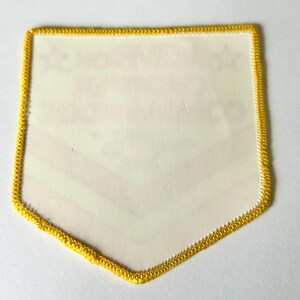 Pu&ograve; includere: Una toppa a forma di scudo con un bordo ricamato giallo brillante. Il centro della toppa &egrave; bianco sporco e presenta testi ed elementi grafici leggermente visibili che sembrano stampati sulla superficie, tra cui due piccole icone a stella.