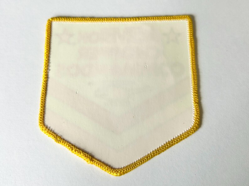 Pu&ograve; includere: Una toppa a forma di scudo con un bordo ricamato giallo brillante. Il centro della toppa &egrave; bianco sporco e presenta testi ed elementi grafici leggermente visibili che sembrano stampati sulla superficie, tra cui due piccole icone a stella.