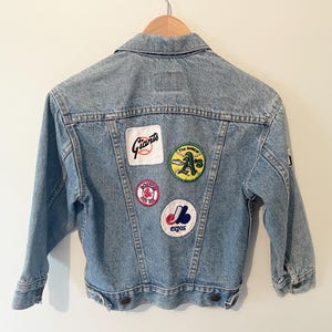 May include: A light blue denim jacket with various baseball team patches on the back, including the Giants, A's, Boston Red Sox, and Expos.