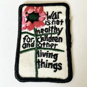 May include: A rectangular embroidered patch with a white background and a thick black border. It features a pink flower with a green center and stem, accompanied by the black embroidered text: War is not healthy for children and other living things.
