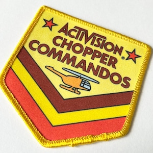 May include: A shield-shaped embroidered patch with a yellow border. The design features the text ACTIVISION CHOPPER COMMANDOS in brown, two red stars, and an orange helicopter graphic above yellow, brown, and red chevron stripes on a yellow background.