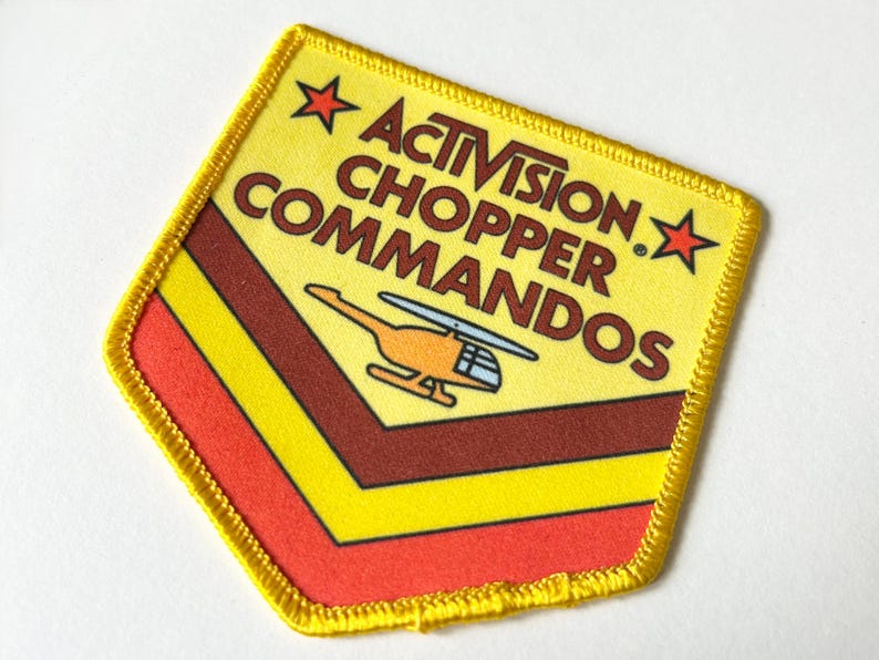 Pu&ograve; includere: Una toppa ricamata a forma di scudo con bordo giallo. Il design presenta il testo ACTIVISION CHOPPER COMMANDOS in marrone, due stelle rosse e un'illustrazione di un elicottero arancione sopra strisce a chevron gialle, marroni e rosse su sfondo giallo.