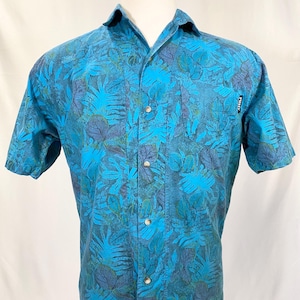 Pode incluir: Uma camisa azul de manga curta com botões e estampa de folhas tropicais. A camisa tem um bolso do lado esquerdo com a palavra "Purity" impressa nele.