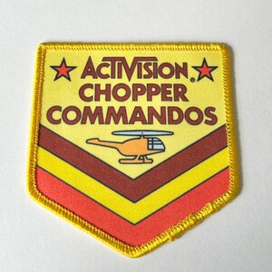Pu&ograve; includere: Una toppa ricamata a forma di scudo con sfondo giallo, che riporta il testo ACTIVISION CHOPPER COMMANDOS in marrone, due stelle rosse e un grafico di un elicottero arancione. La parte inferiore presenta strisce a chevron marroni e rosse.