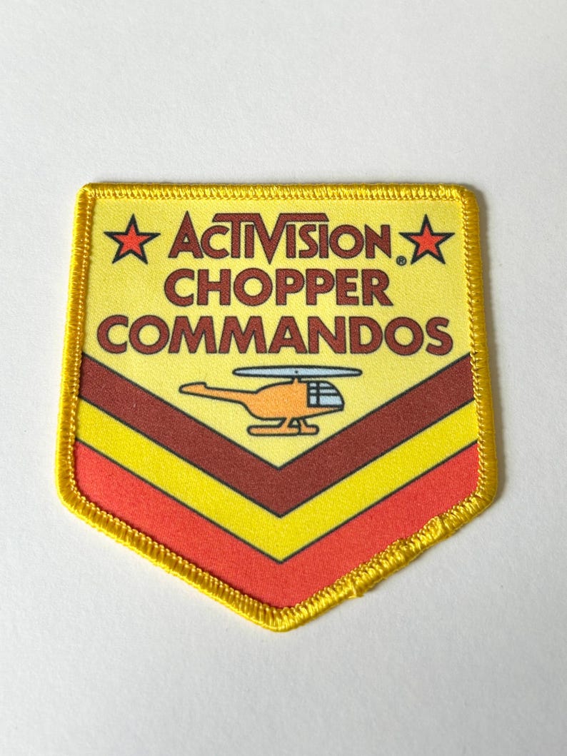 Pu&ograve; includere: Una toppa ricamata a forma di scudo con sfondo giallo, che riporta il testo ACTIVISION CHOPPER COMMANDOS in marrone, due stelle rosse e un grafico di un elicottero arancione. La parte inferiore presenta strisce a chevron marroni e rosse.
