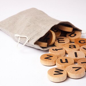Puede incluir: Una bolsa de lino beige con un cord&oacute;n, derramando fichas de letras de madera. Las fichas son circulares, con letras min&uacute;sculas negras. Las letras est&aacute;n esparcidas sobre una superficie blanca, lo que sugiere un juguete educativo.