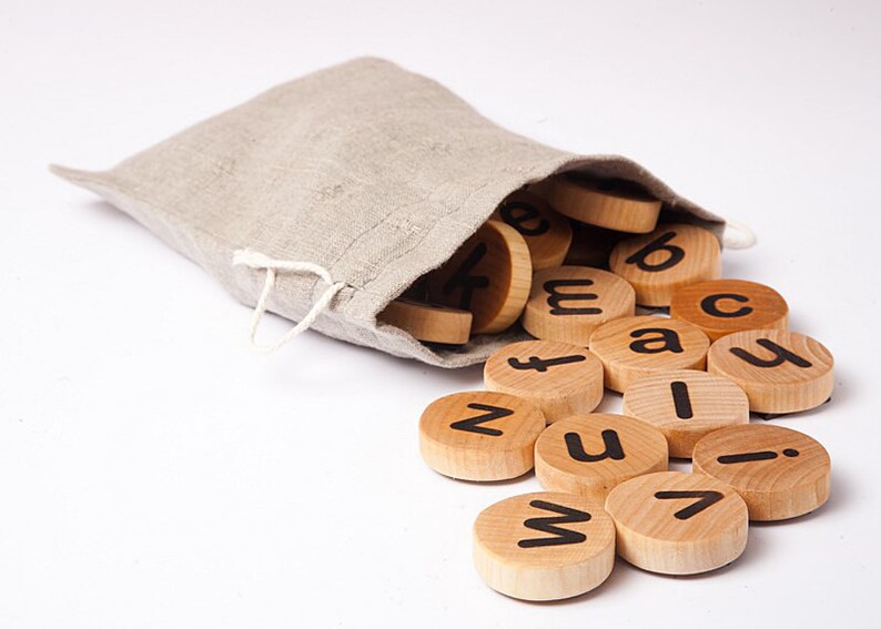 Puede incluir: Una bolsa de lino beige con un cord&oacute;n, derramando fichas de letras de madera. Las fichas son circulares, con letras min&uacute;sculas negras. Las letras est&aacute;n esparcidas sobre una superficie blanca, lo que sugiere un juguete educativo.