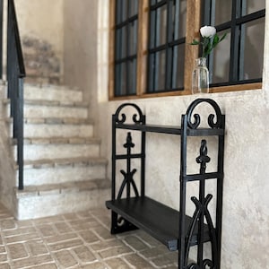 May include: Black metal shelf with two levels, featuring ornate scrollwork and a decorative finial. The shelf is positioned in front of a window with black frames and a staircase in the background. A white rose in a glass vase sits on the windowsill.